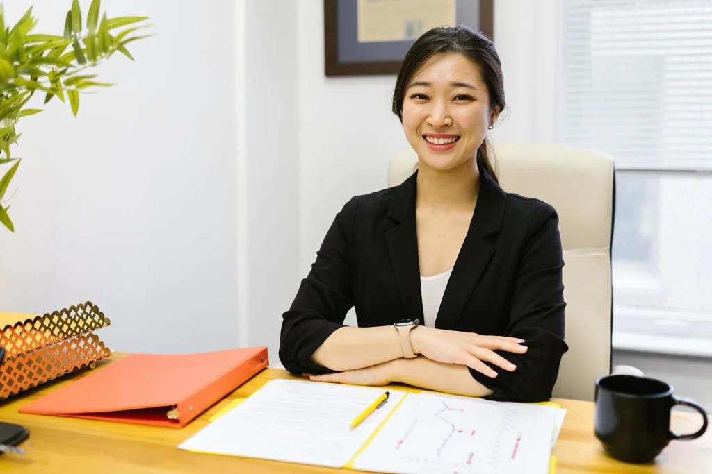 Persoană care stă la un birou zâmbind și privind la aparatul de fotografiat