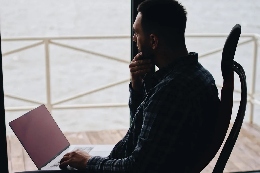Dizüstü bilgisayarda çalışırken pencereden suya bakan kişi