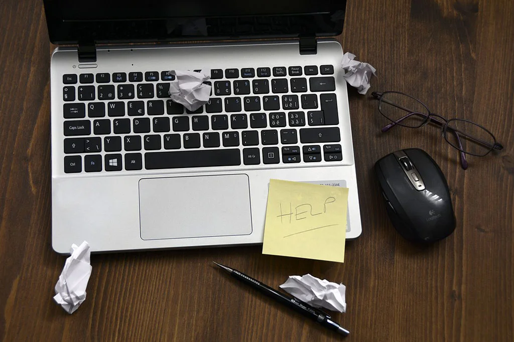 un computer pe un birou cu hârtie prăbușită pe el și un bilețel pe care scrie AJUTOR