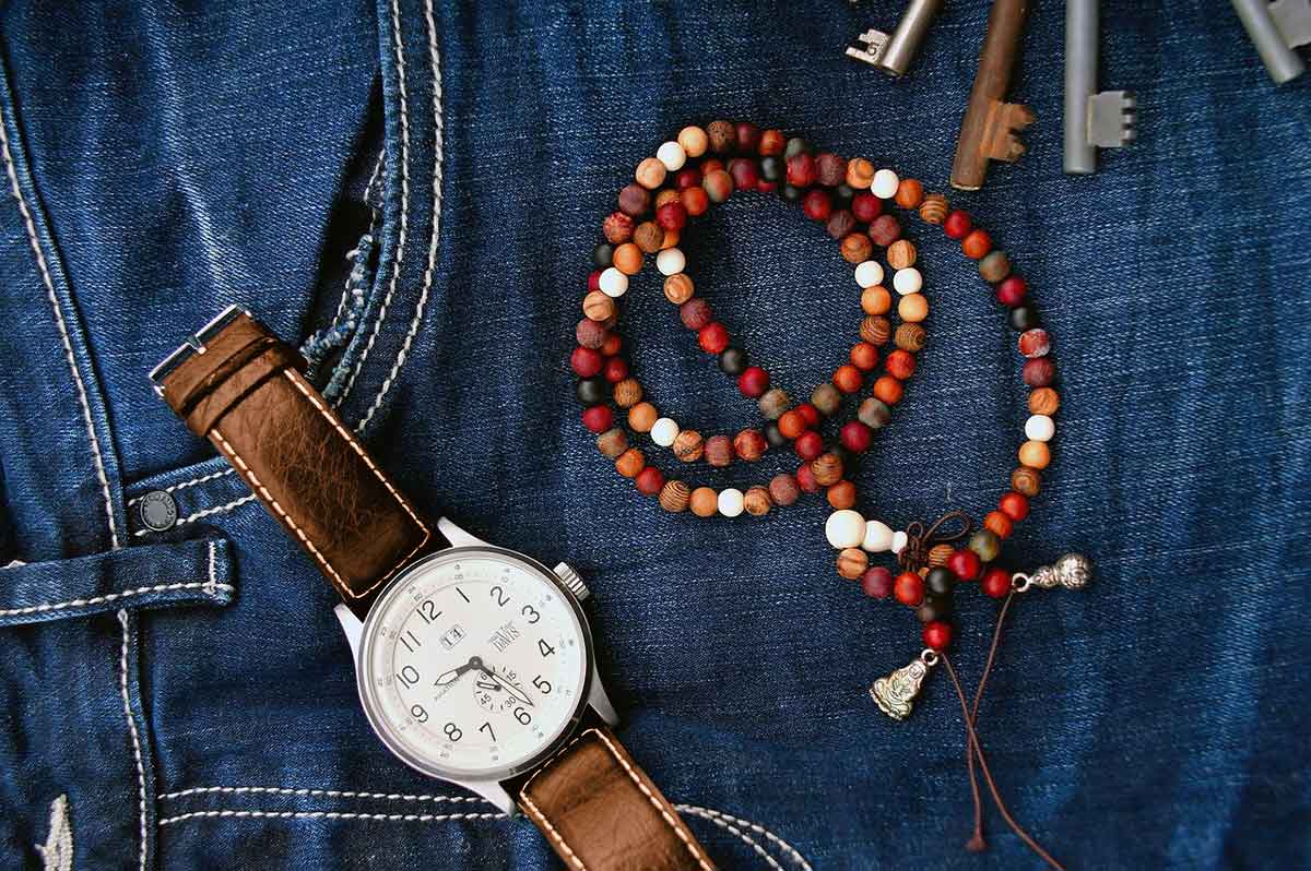 Reloj de cuero marrón y pulsera de cuentas de madera roja, naranja y marrón sobre jeans azules.