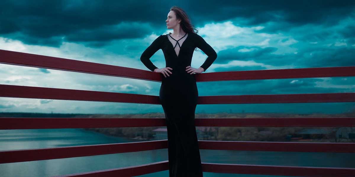 женщина в элегантном длинном черном платье стоит перед темным угрюмым небом.