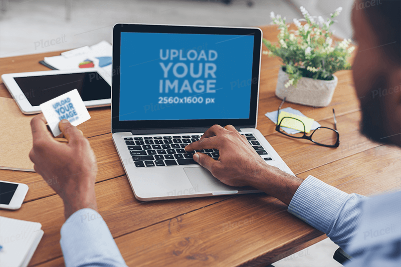 Über der Schulter Mockup eines Mannes, der an seinem Macbook arbeitet und eine Visitenkarte hält